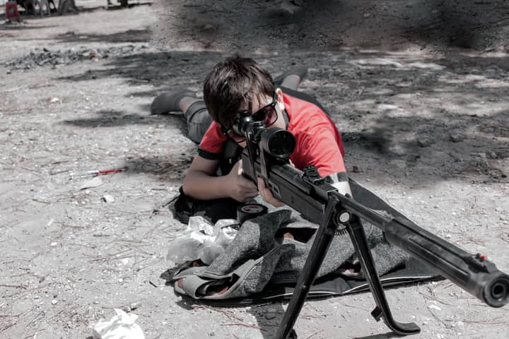 A child is aiming a rifle outdoors, focused and wearing sunglasses, in a realistic training environment.