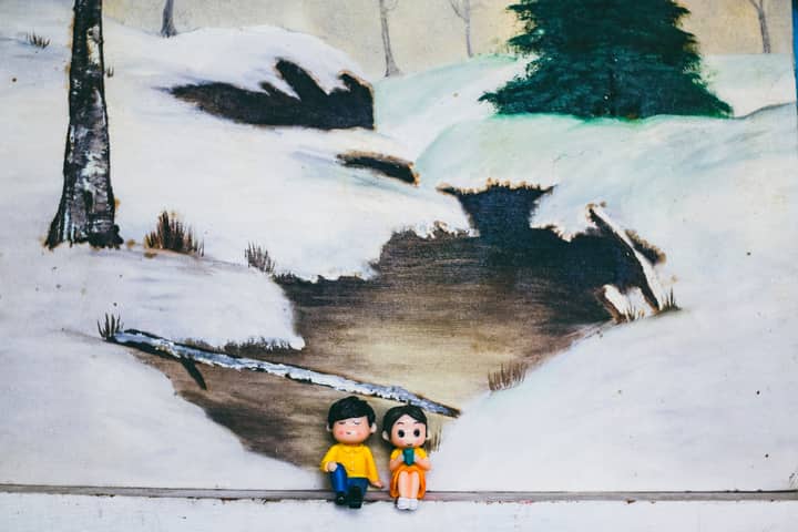 Figures of woman with boyfriend sitting against canvas with painting of river between snowy hills in countryside