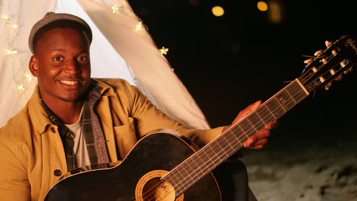 A smiling man plays guitar near a string-lit tent at night, radiating happiness.