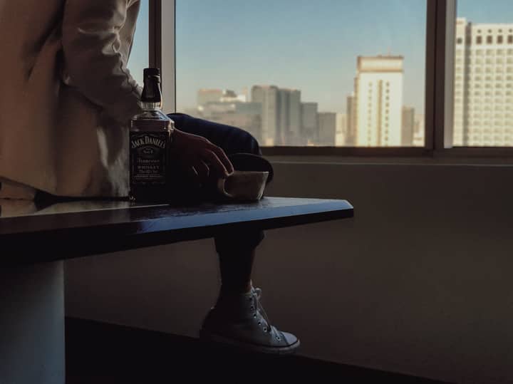 Adult sitting indoors holding a cup beside a bottle of Jack Daniel's, with urban skyline view.