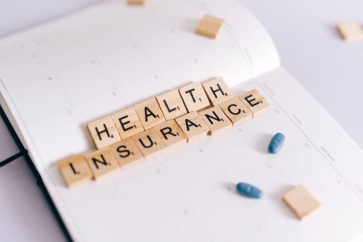 Close-up of health insurance spelled out with Scrabble tiles on a planner, alongside medication pills.