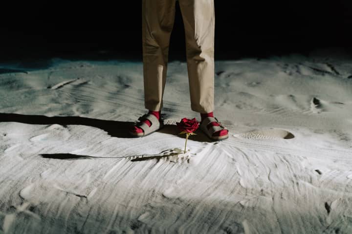 A red rose stands near a person's feet on sandy ground, highlighting contrast and mystery.