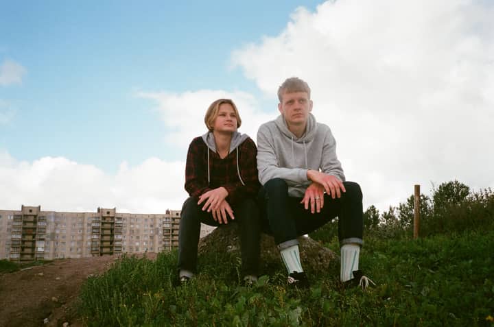 Two young adults sitting on a grass-covered hill with a residential background on a clear day.