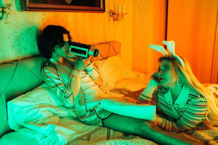 Two adults in a retro-styled bedroom, one with a vintage camera, the other wearing bunny ears.