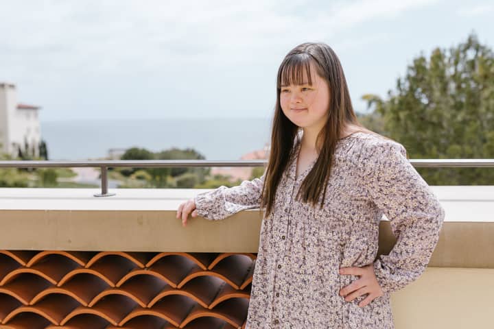 A young woman in a floral top stands outdoors, enjoying a scenic seaside view.