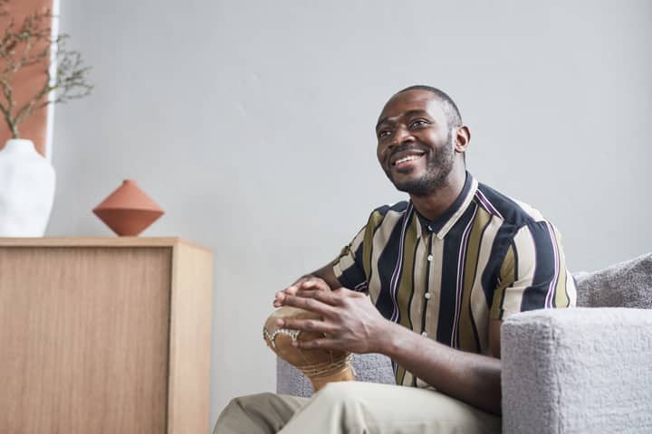 A cheerful African man playing a djembe drum indoors, seated comfortably and smiling.