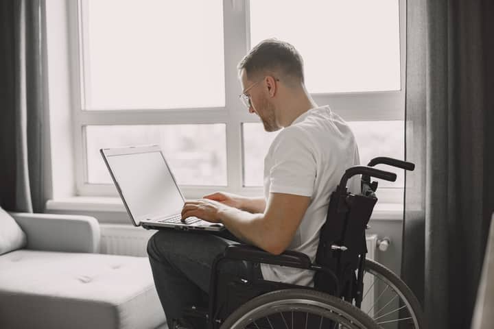 A man in a wheelchair is using a laptop in a bright room with a large window.