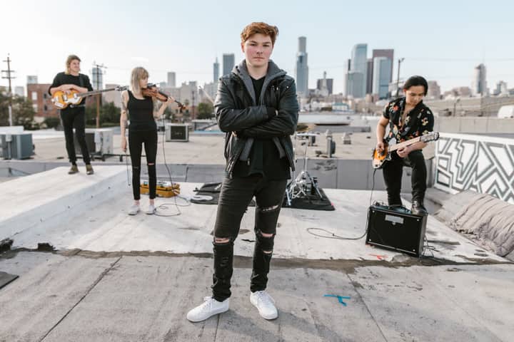 A band performing on an urban rooftop with a city skyline backdrop, featuring guitars and a violin.