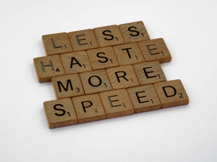 Wooden scrabble tiles forming the phrase 'Less Haste More Speed' on a white background.