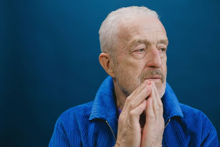 Elderly man with white hair and beard in a blue jacket, hands together, against a blue background.