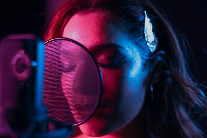 Close-up of a woman singing with eyes closed in a vibrant lit recording studio.