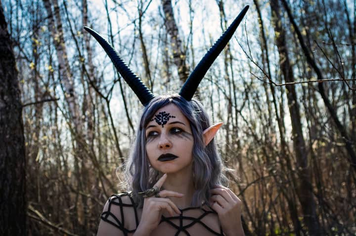 Portrait of young woman with black makeup and purple hair and black horns looking away in forest