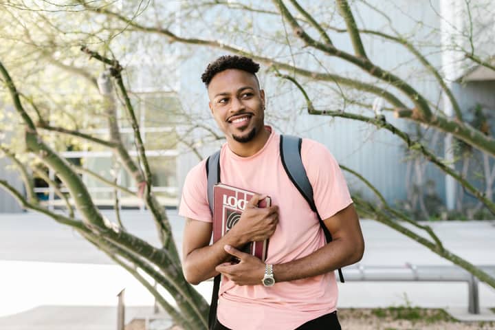 Smiling college student holding books outdoors, wearing a pink t-shirt and backpack.