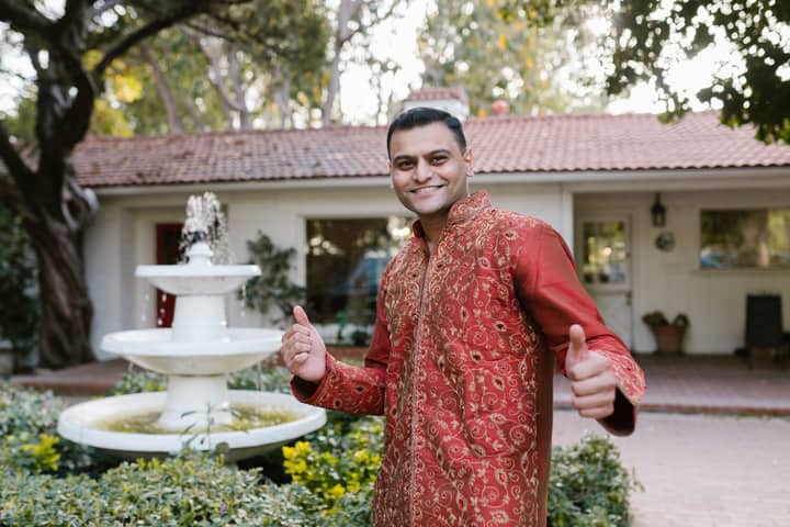 Man in red traditional Indian attire standing outdoors with a fountain in the background, giving thumbs up.
