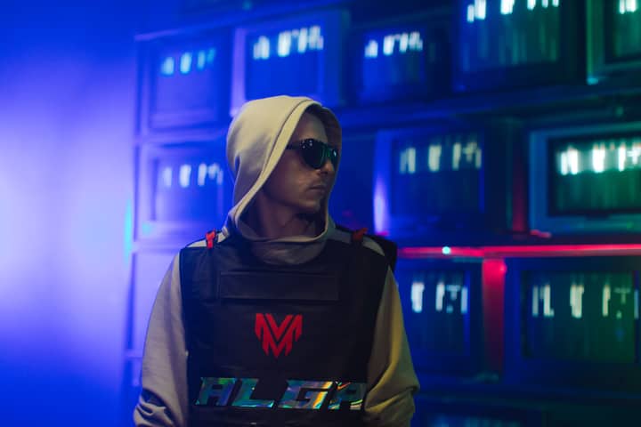 Portrait of a man in a hoodie and sunglasses, with a neon-lit background of screens.