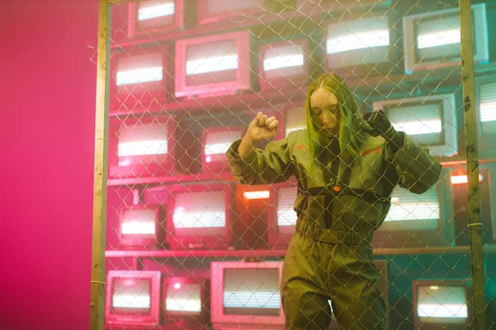 A woman in a jumpsuit stands behind a wire fence in a room filled with glowing TV screens.