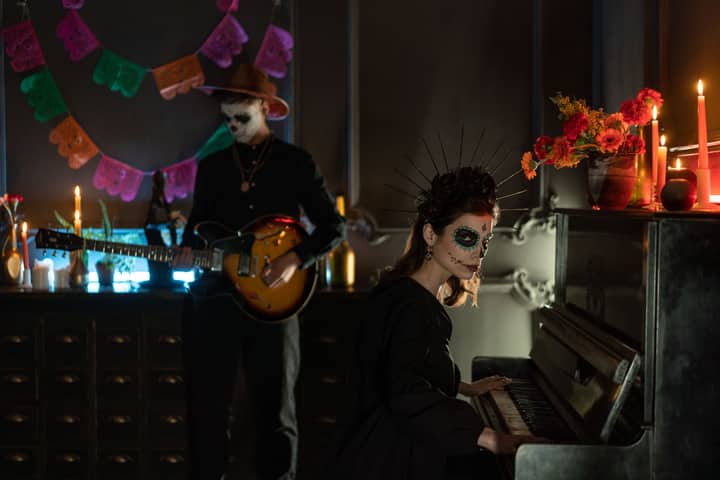 Musicians celebrate Dia de los Muertos with traditional face paint, playing guitar and piano indoors.