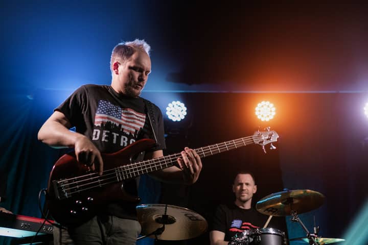 Two musicians performing with bass guitar and drums under colorful stage lights.