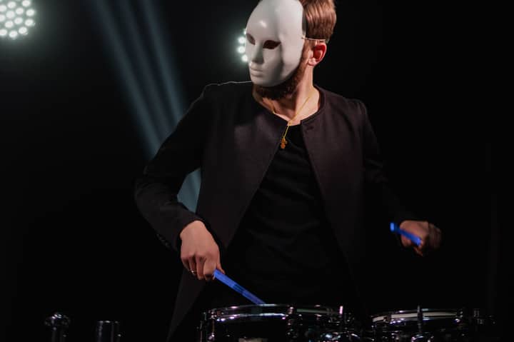 A drummer wearing a white mask plays energetically on stage under spotlights, creating a mysterious atmosphere.