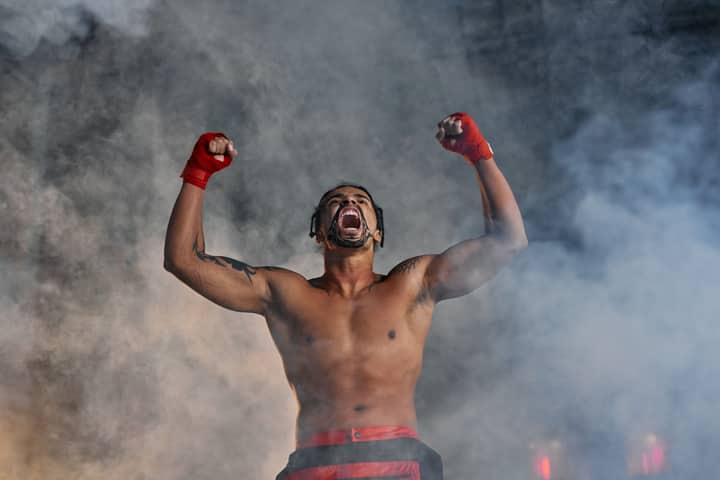 Shirtless fighter shouting with raised fists amidst smoke, portraying strength and intensity.