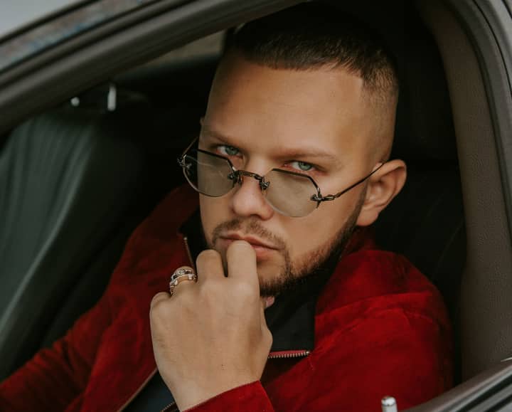 Pensive young unshaven male in eyeglasses and red jacket sitting on driver front seat of car and looking at camera
