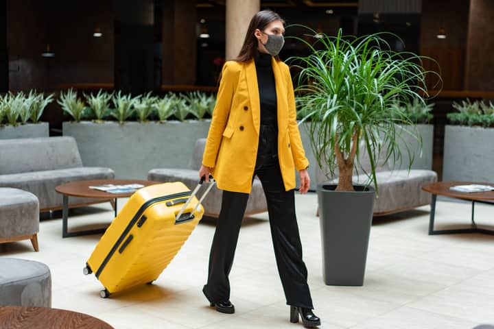 Fashionable woman in yellow blazer and face mask walking with suitcase indoors.