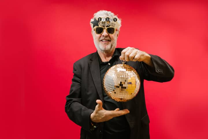 A cheerful elderly man with sunglasses and a disco ball against a vivid red background.