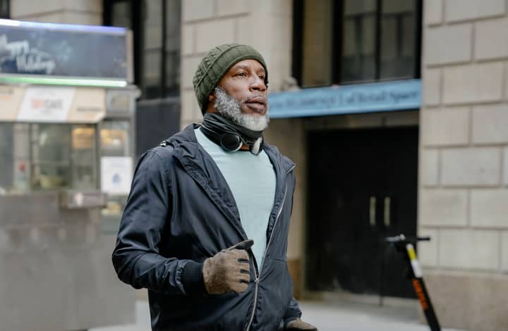 Serious African American bearded male in knitted hat and windbreaker jacket and headphones on neck running in street