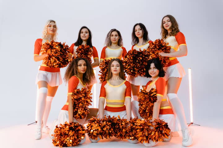 Group of diverse cheerleaders in vibrant uniforms posing with pompoms in a studio setting.