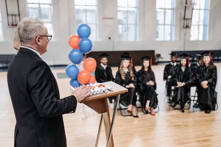A formal graduation ceremony with a speaker addressing seated graduates in a gymnasium.