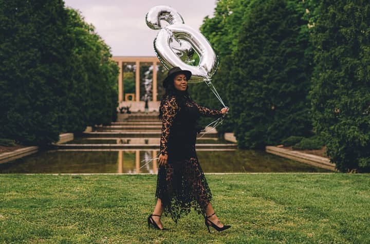 Full body side view of African American female in hat looking at camera while strolling near pond with foil balloons in shape of number 30 during birthday celebration