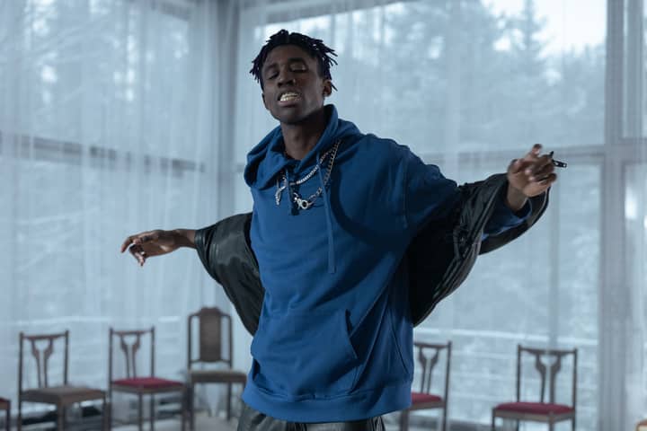 Black rapper energetically performing indoors in a blue hoodie and silver chains, conveying passion and rhythm.