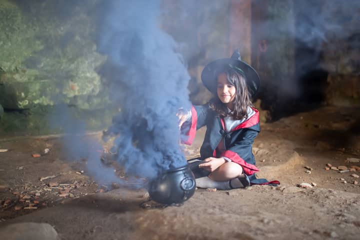 A young girl dressed as a witch sitting with a smoking cauldron, creating a magical scene.
