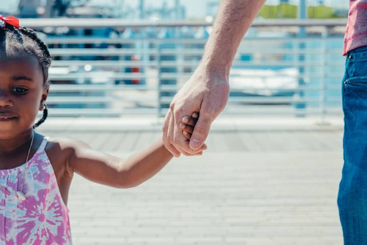 A close-up of a father holding hands with his young daughter outdoors on a sunny day.