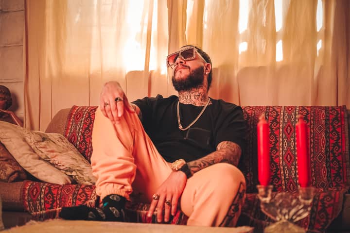 Bearded man with tattoos and sunglasses sits relaxed on a stylish sofa inside a cozy room.