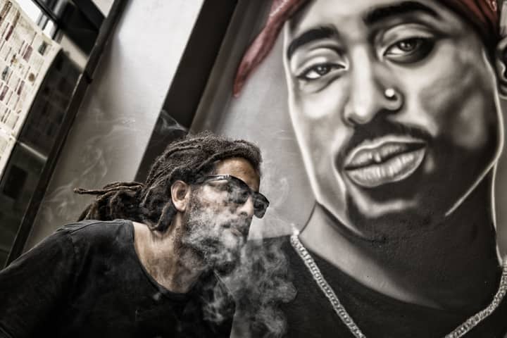 A man smoking in front of a mural featuring a hip hop artist, blending urban style and art.