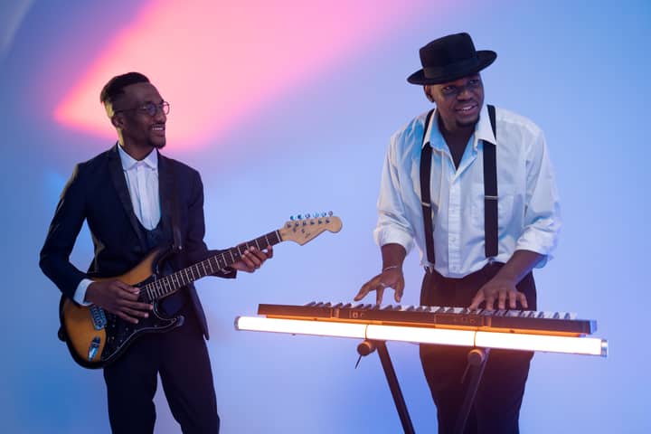 Two musicians in vibrant lighting play electric guitar and keyboard indoors.