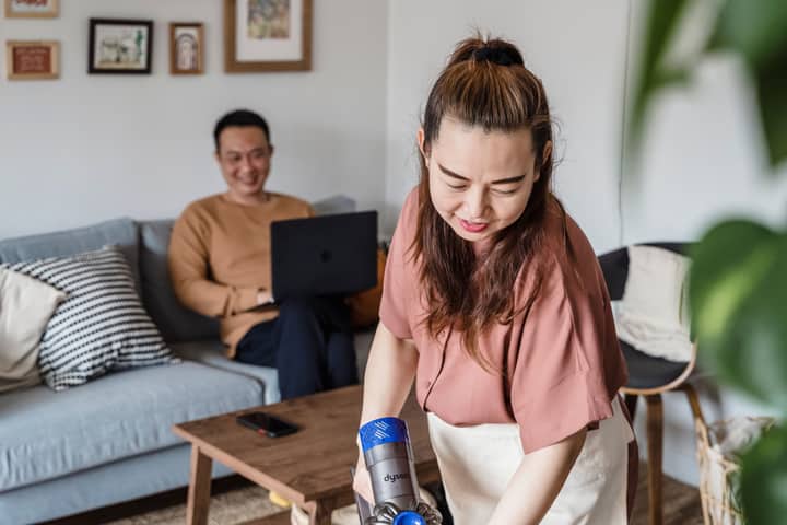A woman smiling while vacuuming a living room, with a man working on a laptop in the background.