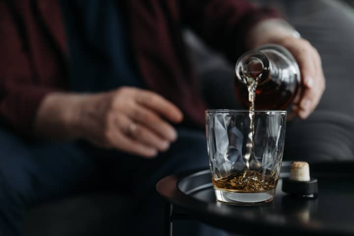 An adult pours whiskey from a bottle into a glass on a table. Indoors setting.