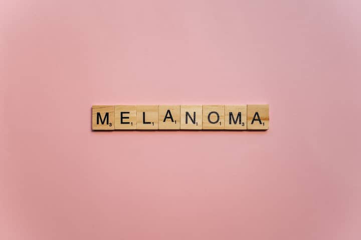 Wooden Scrabble tiles spelling 'Melanoma' on a soft pink background, symbolizing awareness.