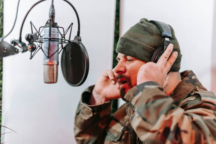 Man in a camouflage jacket singing into a microphone in a studio setting with headphones on, focused and expressive.