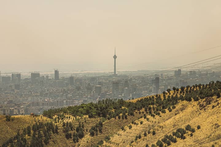 View of Tehran cityscape featuring the Milad Tower surrounded by hills under hazy skies.