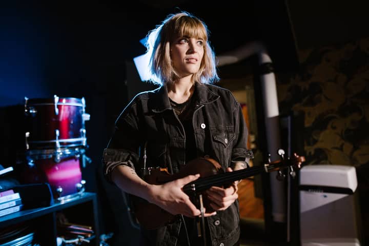 A young caucasian woman holding a violin in a dimly lit music studio, looking away thoughtfully.