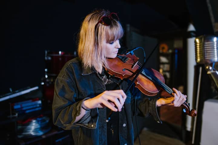 Female violinist performing indoors, wearing a denim jacket, with vintage microphone nearby.