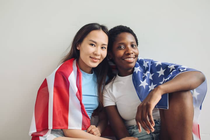 Two diverse women celebrating Independence Day with an American flag. Smiling and sitting indoors.