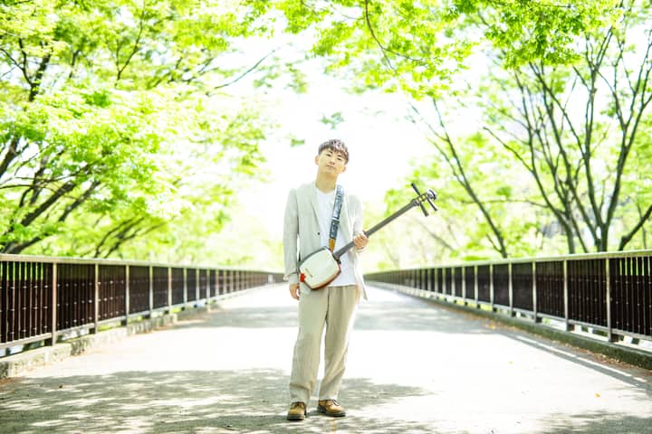 An Asian man standing on a sunlit bridge, holding a shamisen, surrounded by vibrant green trees.
