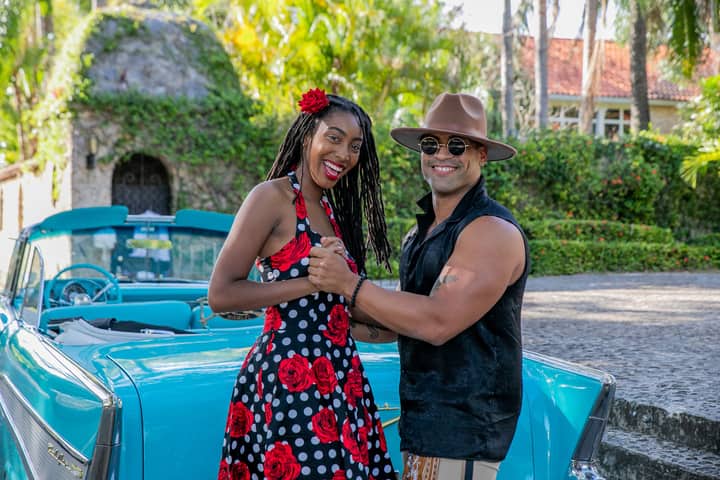 Smiling couple dances joyfully by a turquoise classic car in a lush outdoor setting.