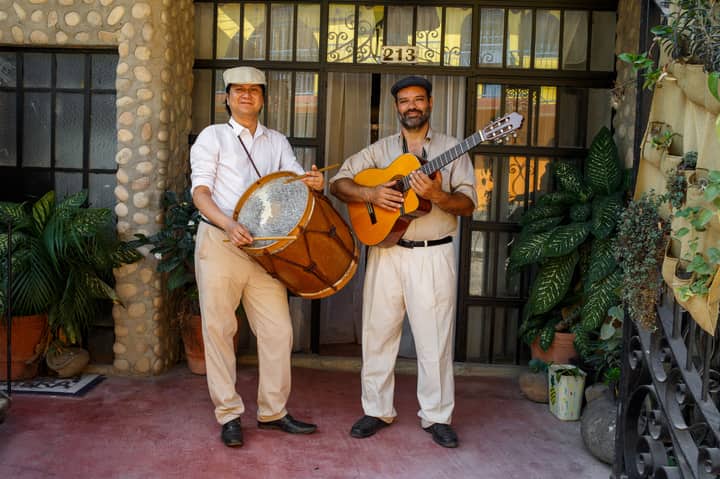 Two Hispanic musicians playing a guitar and a drum, posing outdoors in a traditional setting.