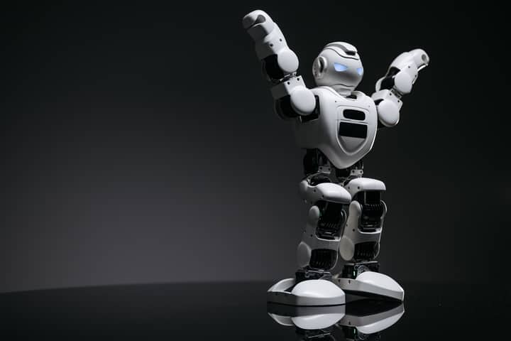 Close-up studio shot of a white robot toy with LED eyes raised in victory on a gray background.