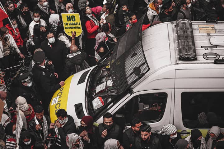 A crowd of protesters surrounding a police van holding signs during a demonstration against Israeli occupation.